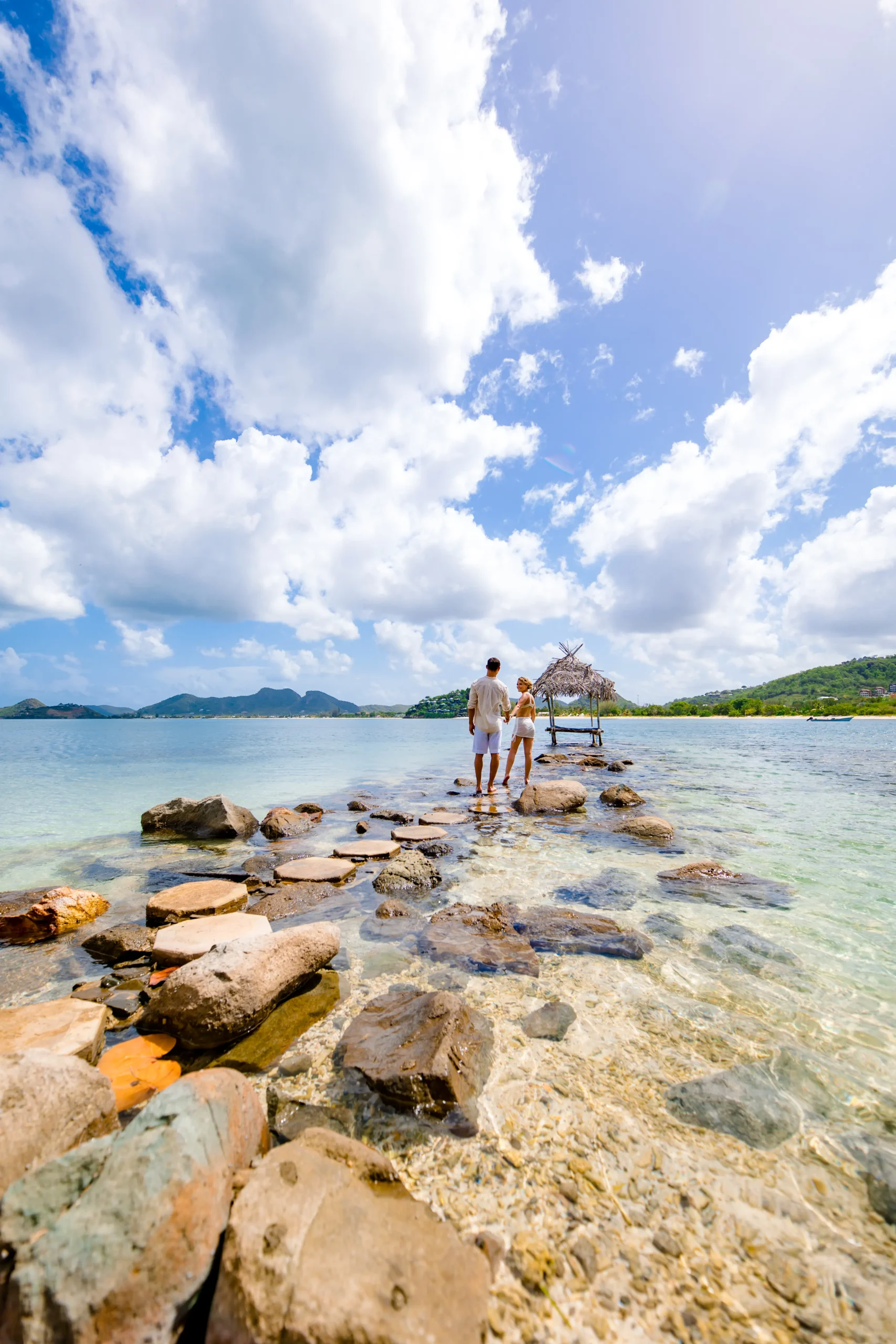 Couple walking hand in hand along a stone jetty over clear turquoise water in Antigua with green hills and a thatched gazebo in the background